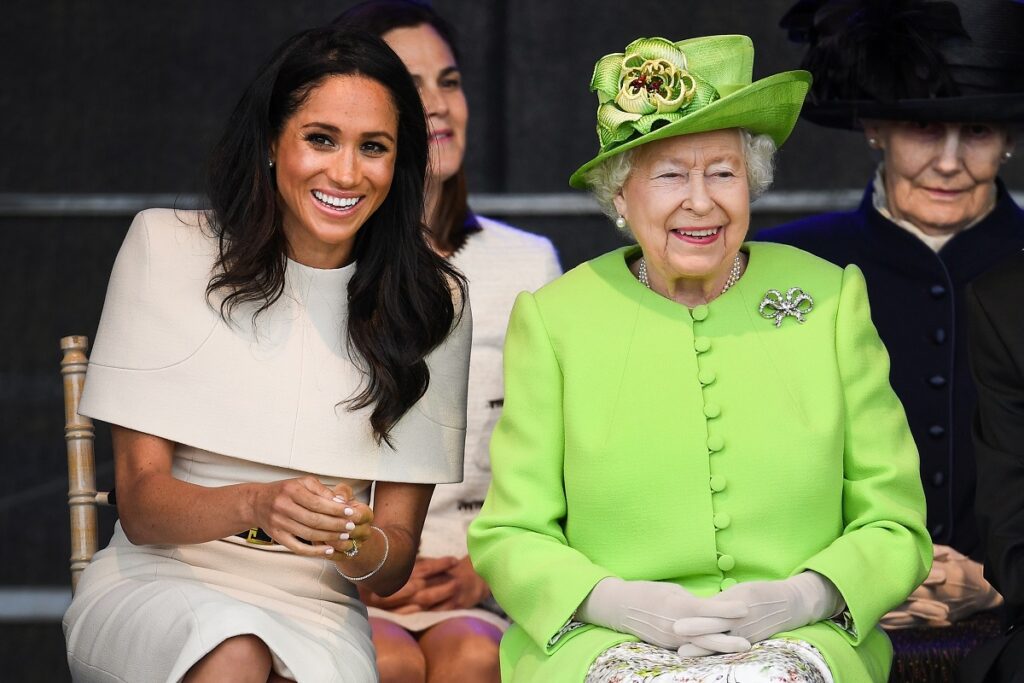 Meghan Markle îmbrăcată într-o rochie albă în timp ce zâmbește alături de Regina Elisabeta care a cunoscut-o pe fiica Ducilor de Sussex printr-o conferință video, în timp ce participă la ceremonia de deschidere a podului de la Widnes în iunie 2018