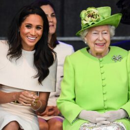 Meghan Markle îmbrăcată într-o rochie albă în timp ce zâmbește alături de Regina Elisabeta care a cunoscut-o pe fiica Ducilor de Sussex printr-o conferință video, în timp ce participă la ceremonia de deschidere a podului de la Widnes în iunie 2018