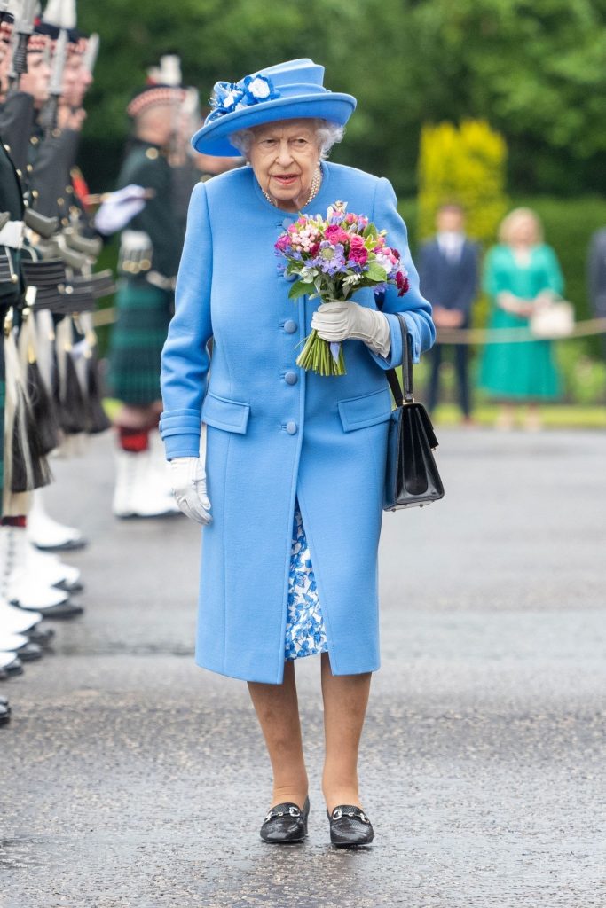 Regina Elisabeta într-un costum albastru și o pălărie de aceasși culoare în timp ce este întâmpinată în mod oficial la vizita sa din Scoția