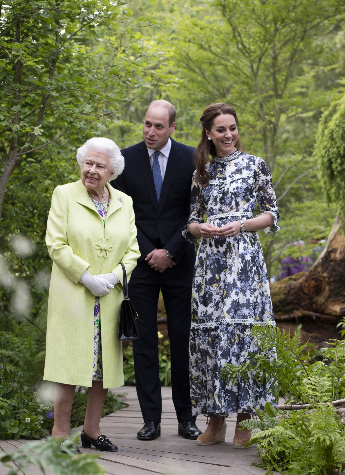 Regina Elisabeta purtând un costum galben alături de Prințul William la costum negru și Ducesa de Cambridge, Kate Middleton, îmbrăcată într-o rochie albă cu imprimeu floral în timp ce se plimbă pe aleea unei grădini botanice