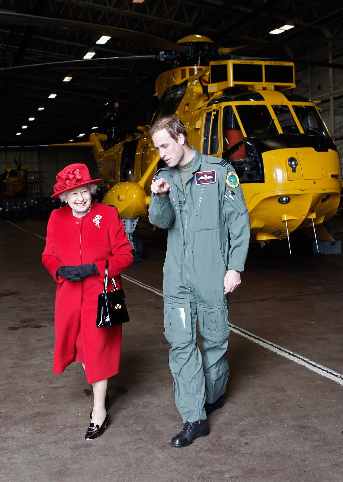 Regina Elisabeta îmbrăcată într-un costum roșu și purtând o pălărie de aceeasși culoare în timp ce merge alături de nepotul său, Prințul William care poartă un costum de militar