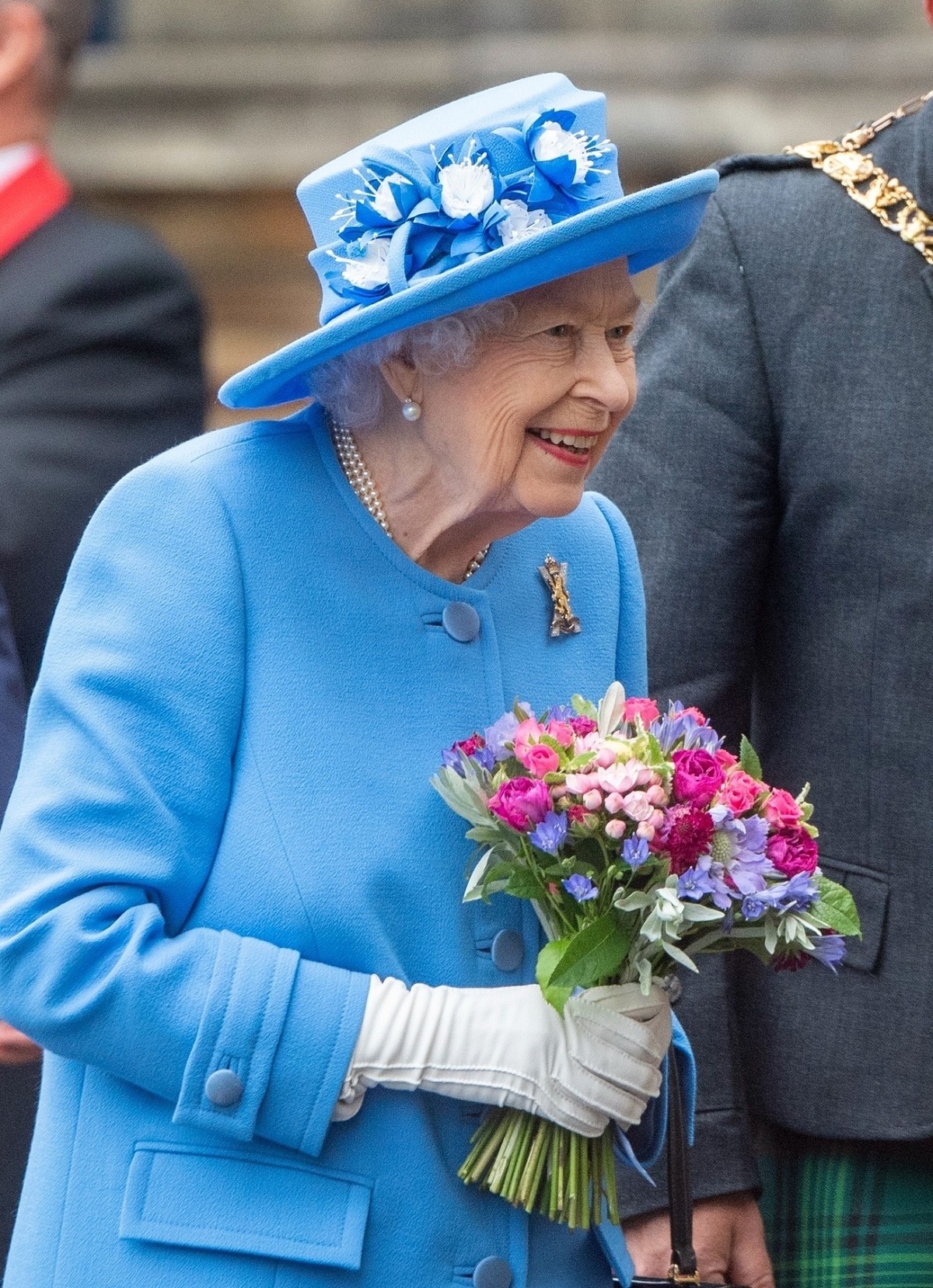 Regina Elisabeta purtând un costum albastru și o pălărie în timp ce merge la vizita unei fabrici de suc din Scoția