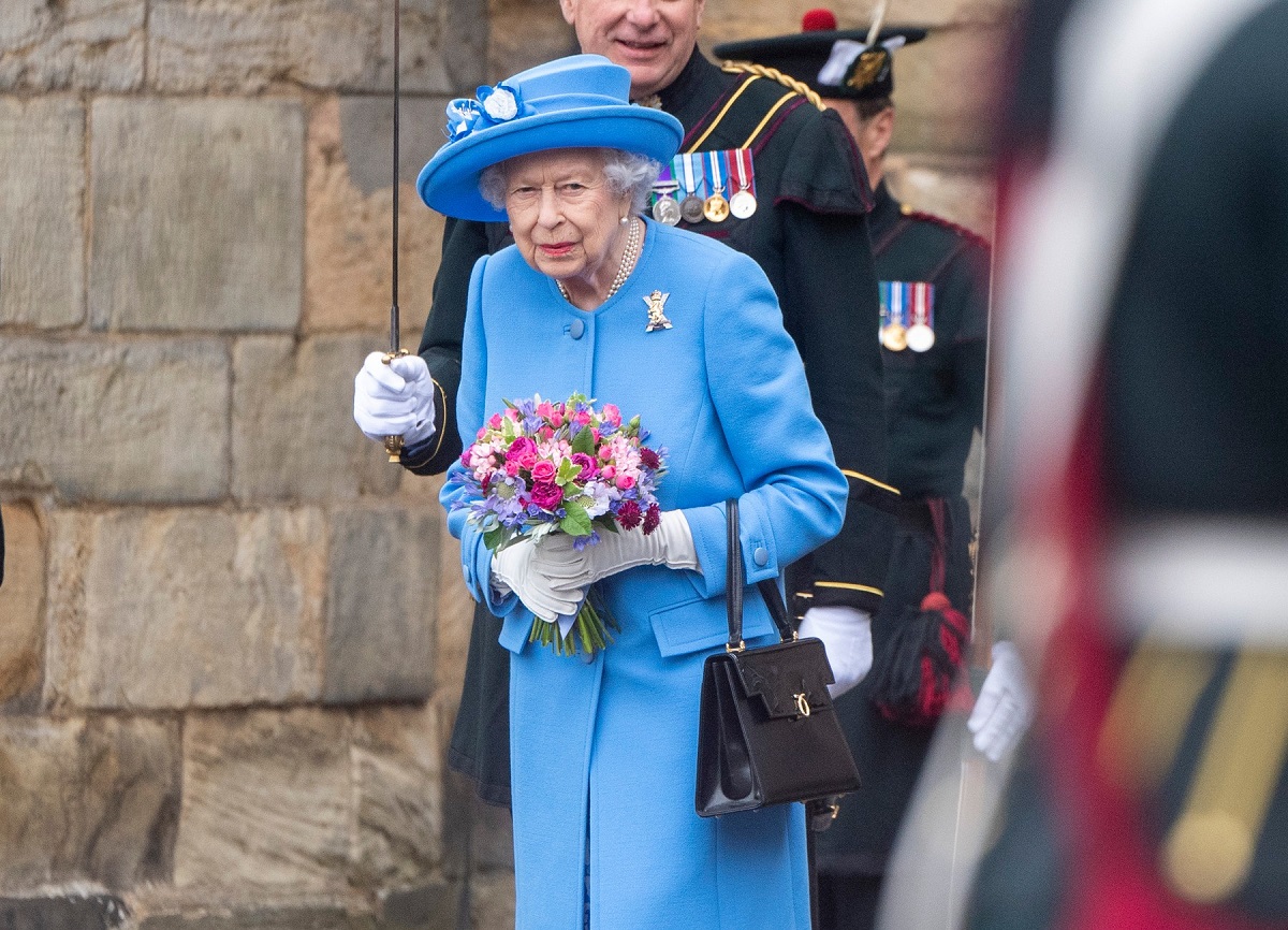 Regina Elisabeta purtând un costum albastru în timp ce ține în mână un buchet de flori la vizita sa oficială de la o fabrică de suc din Scoția
