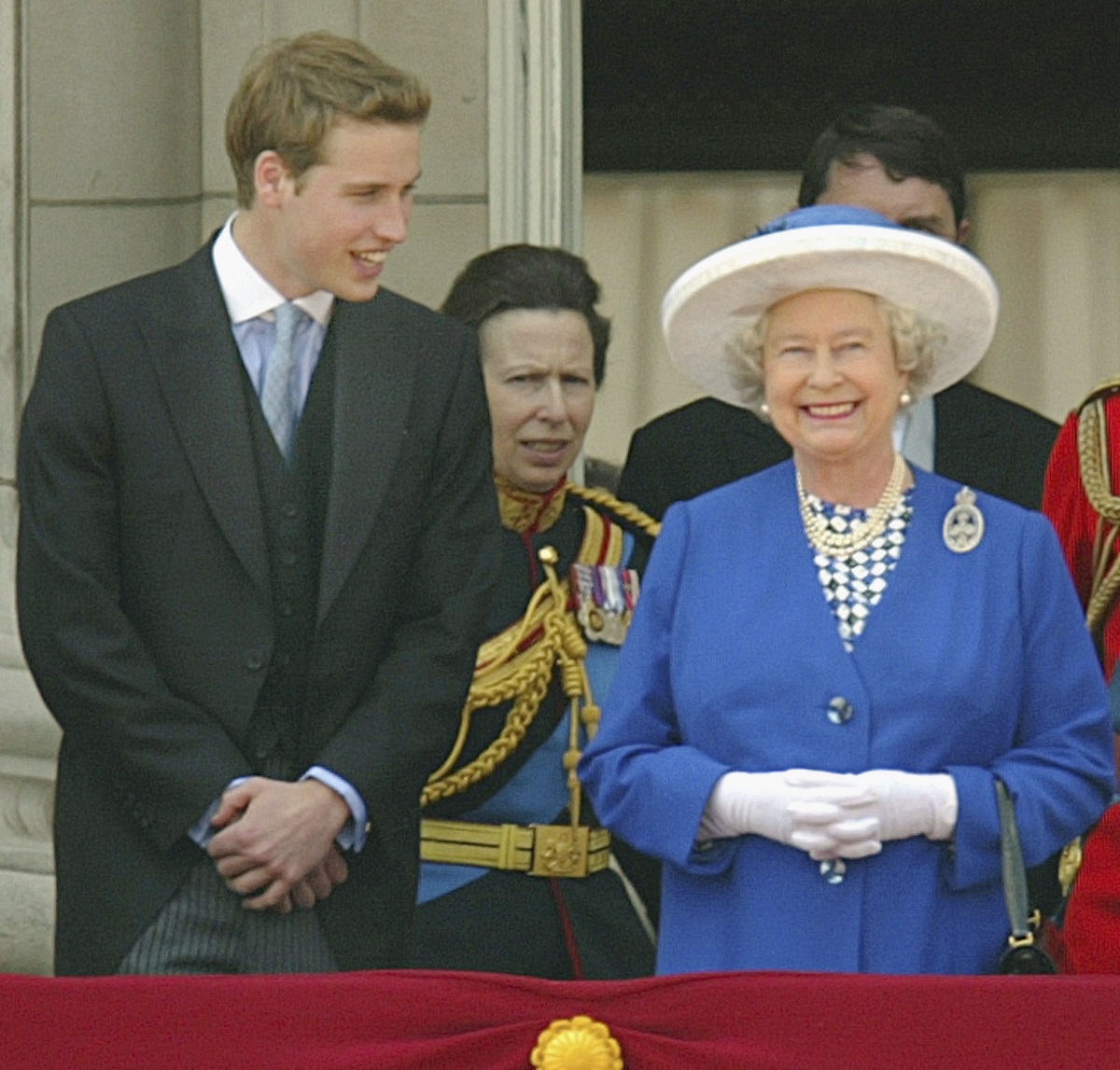 Prințul William la costum negru alături de Regina Elisabeta îmbrăcată în costum albastru și purtând mănuși și pălărie albă în timp ce privesc de la balcon ceremonia Trooping the Colour din 2003