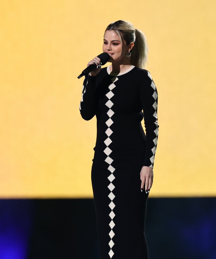 Selena Gomez pe scenă ținând microfonul în mână în timp ce poartă o rochie lungă neagră cu romburi albe și dezvăluie secretul unei ținute perfecte la Global Citizen Vax Live în luna mai 2021