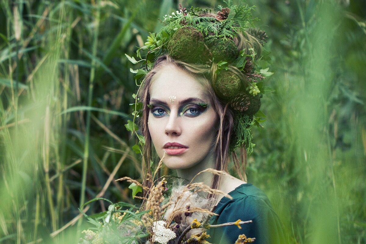 O femeie frumoasă cu machiaj de zână care stă în iarbă și în păr are diferite flori, conuri și crenguțe pentru a reprezenta una din zodiile de pământ