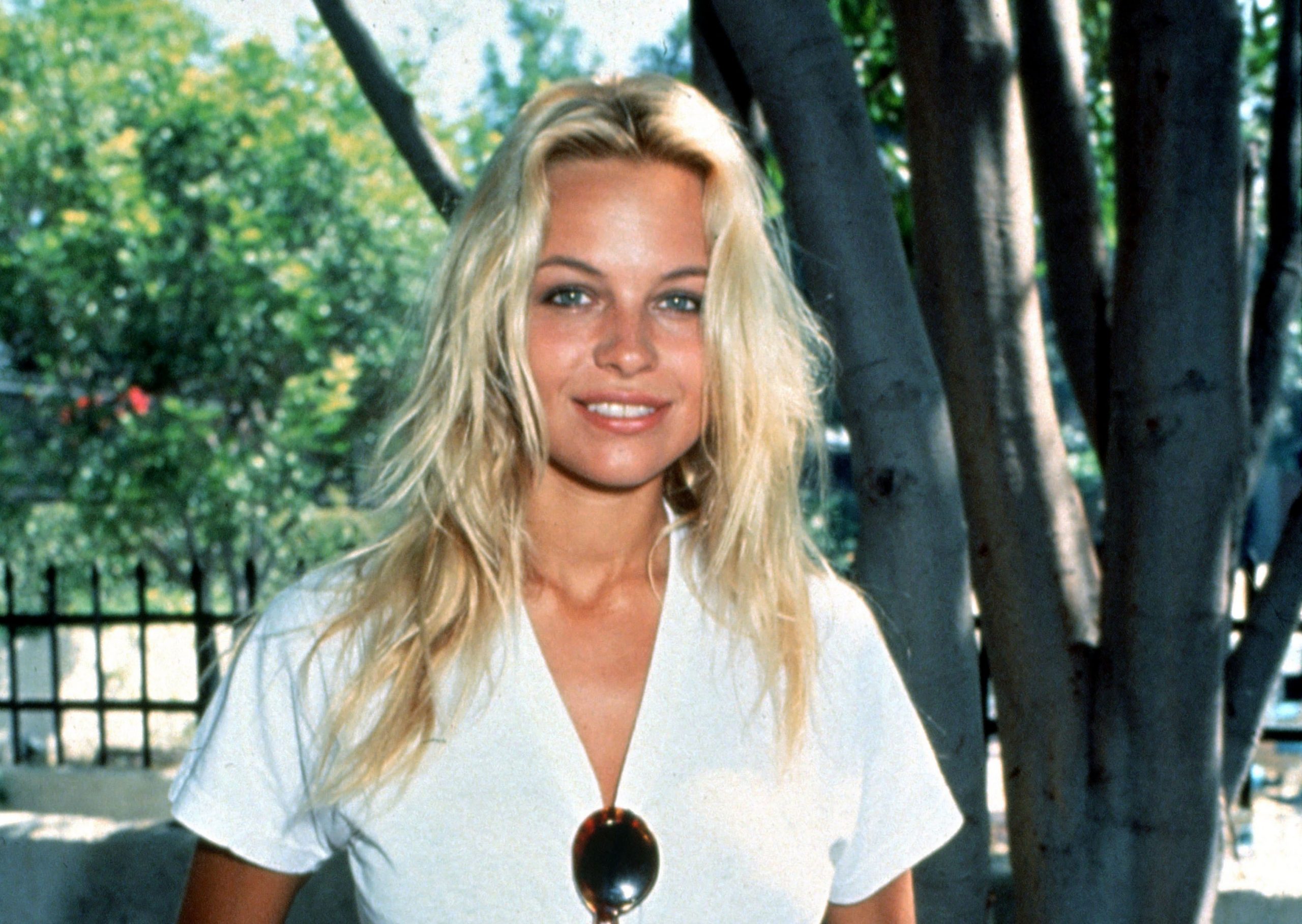 Pamela Anderson avea un look mai natural în tinerețe, optând pentru tricouri. În imagine are un tricou alb, părul lung, ochelari de soare prinși de bluză