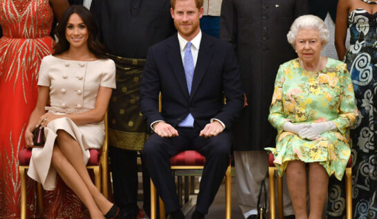 Ducii de Sussex, alături de Regina Elisabeta a II-a, la o ceremonie de premiere, îmbrăcați elegant, pe scaune, în 2018