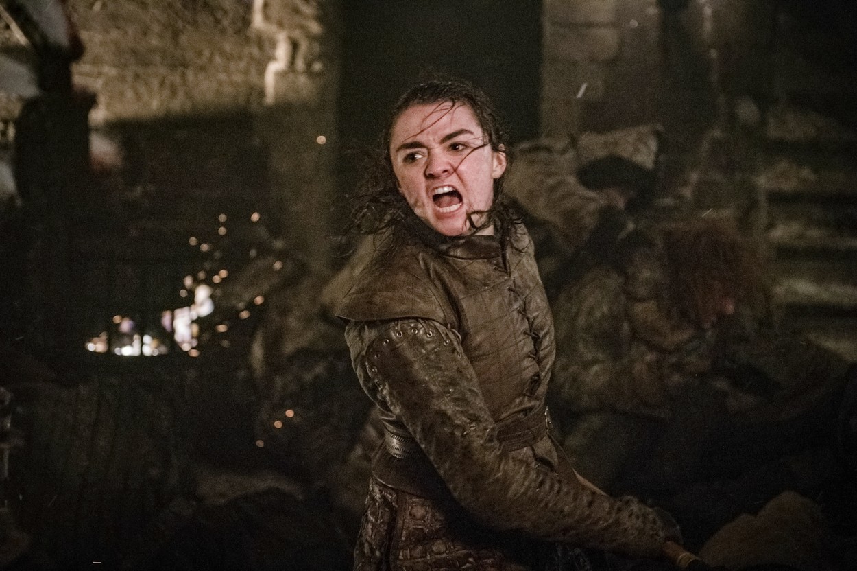 Maisie Williams în rolul lui Arya Stark, Game of Thrones, în timpul unei bătălii din sezonul 8, fundal maro și închis