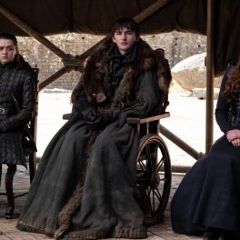 Imagine din sezonul 8 Game of Thrones cu Maisie Williams (Arya Stark), Isaac Hempstead Wright (Bran Stark), Sophie Turner (Sansa Stark), îmbrăcați în negru, pe scaune, fundal deschis la culoare