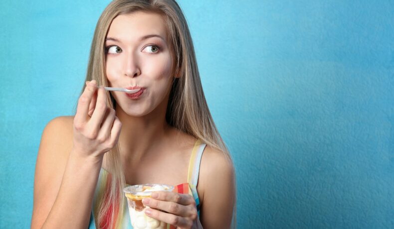 O femeie frumoasă cu părul blond în timp ce stă cu spatele la un perete albastru în timp ce mănâncă un iaurt cu fructe de pădure