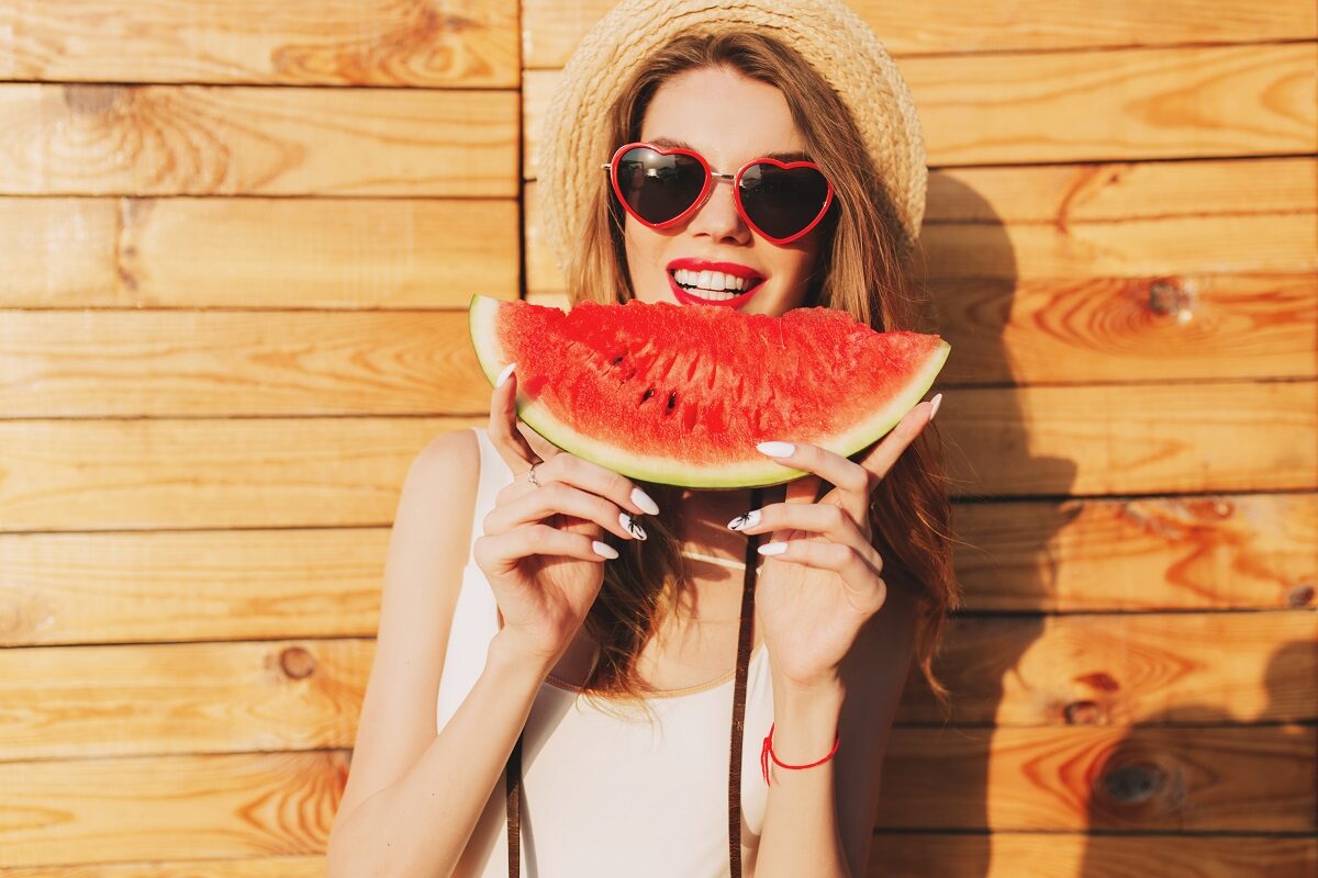 O femeie frumoasă cu ochelari de soare și pălărie de plajă care ține în mâini o felie de pepene și vrea să muște din ea deoarece a aflat care sunt acele beneficii ale consumului de pepene roșu