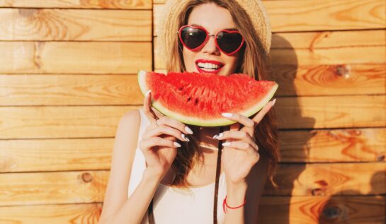 O femeie frumoasă cu ochelari de soare și pălărie de plajă care ține în mâini o felie de pepene și vrea să muște din ea deoarece a aflat care sunt acele beneficii ale consumului de pepene roșu