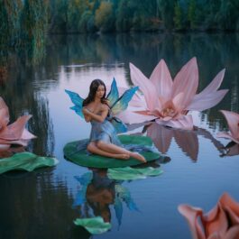 O zână frumoasă cu aripi albastre în timp ce stă pe o frunză de nufăr pe un lac și este înconjurată de flori roz de nufăr