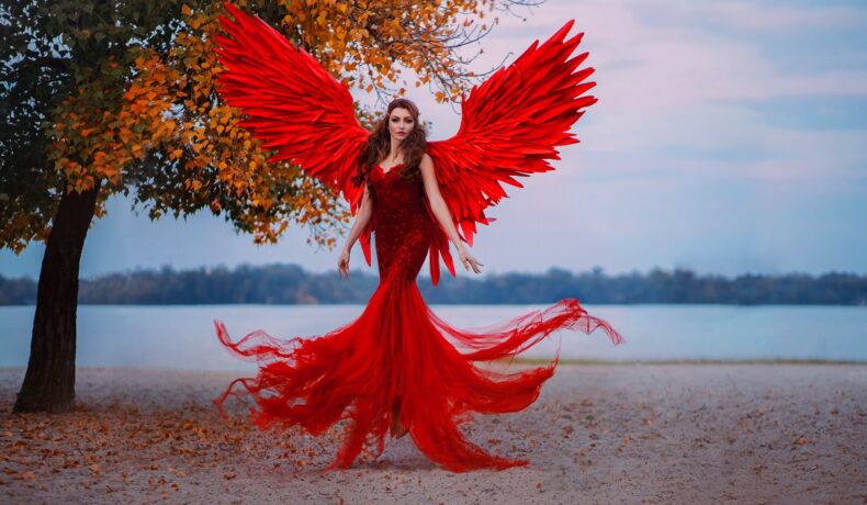 Un cadru feeric din fața unui lac unde se află o femeie superbă cu rochie roșie și aripi de înger roșii pentru a demonstra că este una dintre cele mai pasionale femei din zodiac