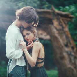 Un cadru feeric dintr-o pădure cu un tânăr în cămașă care o ține pe iubita sa în rochie albastră în brațe și o mângâie pe față