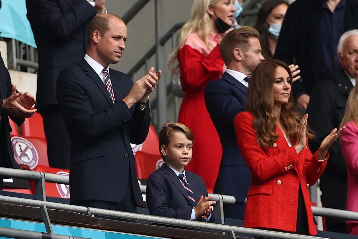 Prințul William alături de Prințul George și Ducesa Kate Middleton într-o tribună la un stadion din Anglia în timp ce aplaudă jucătorii de fotbal