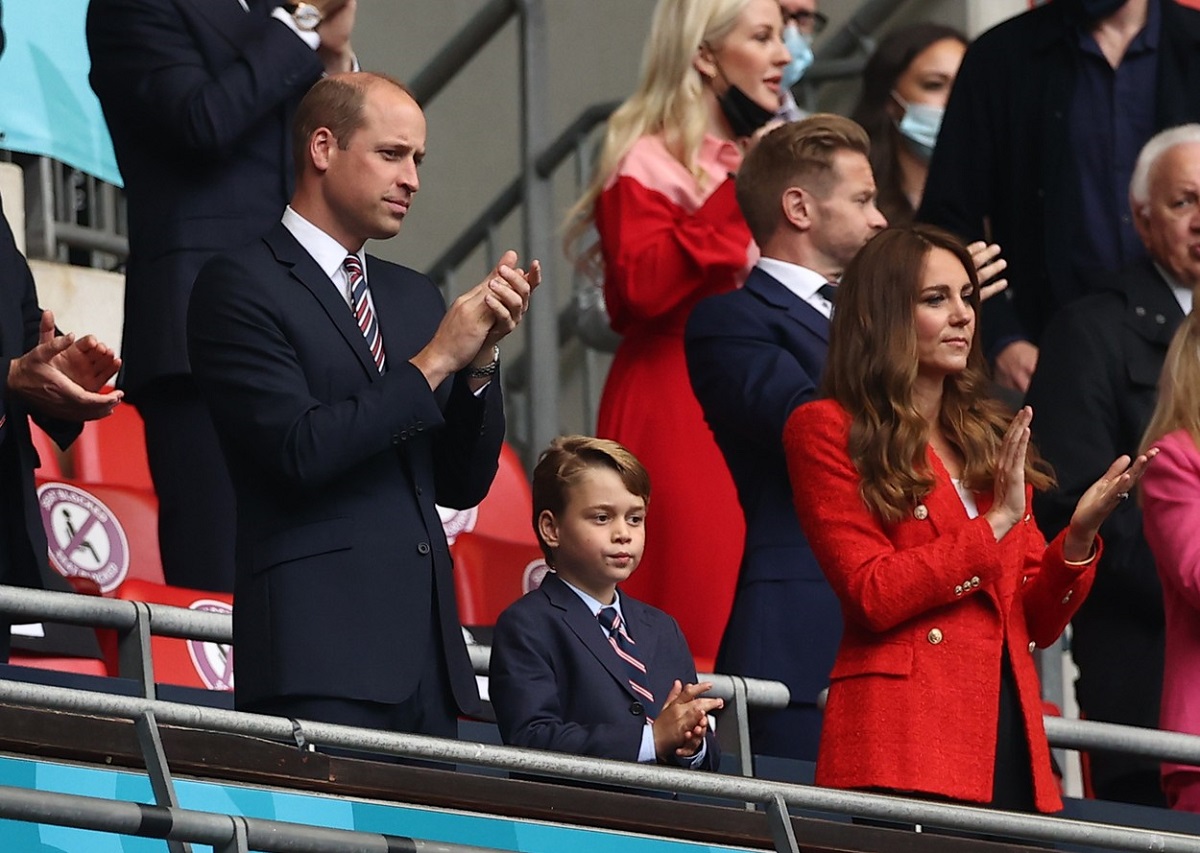 Prințul William alături de Prințul George și Ducesa Kate Middleton într-o tribună la un stadion din Anglia în timp ce aplaudă jucătorii de fotbal