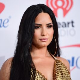 Demi Lovato, pe covorul roșu la Jingle Ball în 2018. Fundal alb, ea e îmbrăcată în rochie aurie, cu părul lung și negru cercei mari