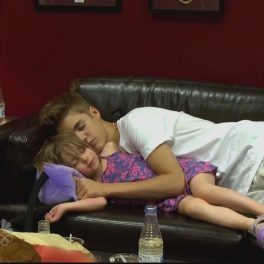 Justin Bieber într-un costum alb de scenă în timp ce doarme pe o canapea și o ține în brațe pe sora lui mai mică în timpul turneului All Around The World
