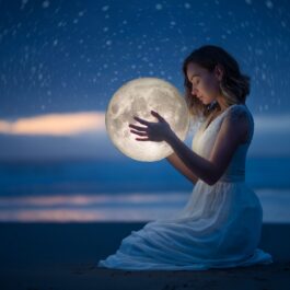 O femeie frumoasă care stă pe plajă în genunchi noaptea în timp ce ține în mâini Luna Plină intrată în zodia Vărsătorilor