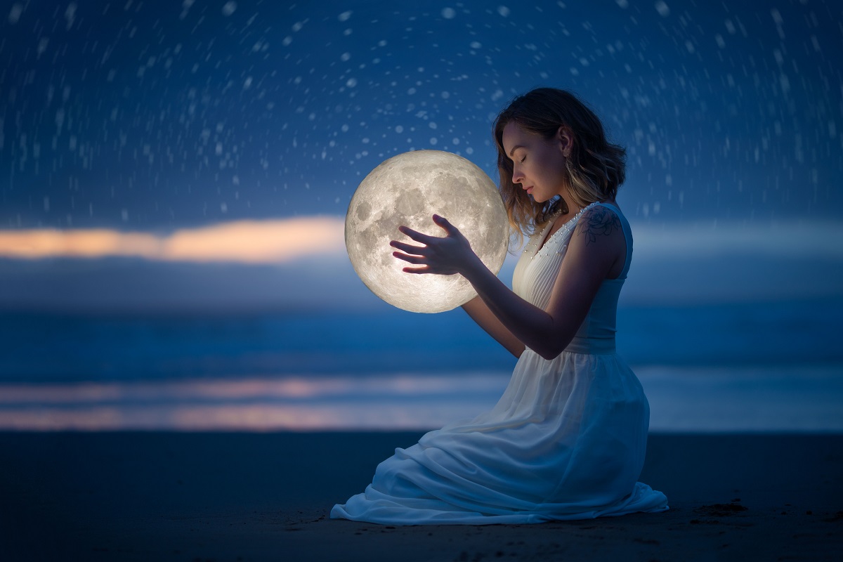 O femeie frumoasă care stă pe plajă în genunchi noaptea în timp ce ține în mâini Luna Plină intrată în zodia Vărsătorilor