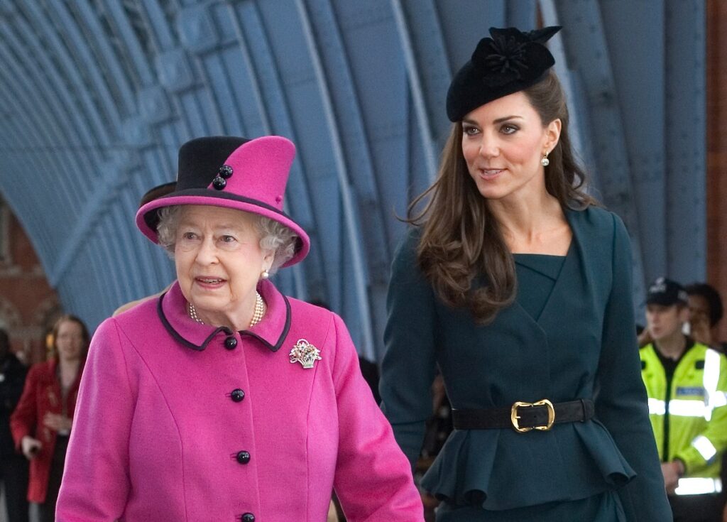 Regina Elisabeta într-un costum roz cu pălărie pe cap alături de Kate Middleton într-un costum verde smarland în timp ce aștaptă un tren pentru a vizita Leicester în 2012