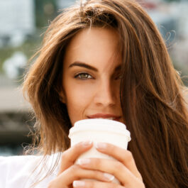 O femeie frumoasă, cu părul lung, ține în mâini un pahar alb de unică folosință, în care se află cafea.