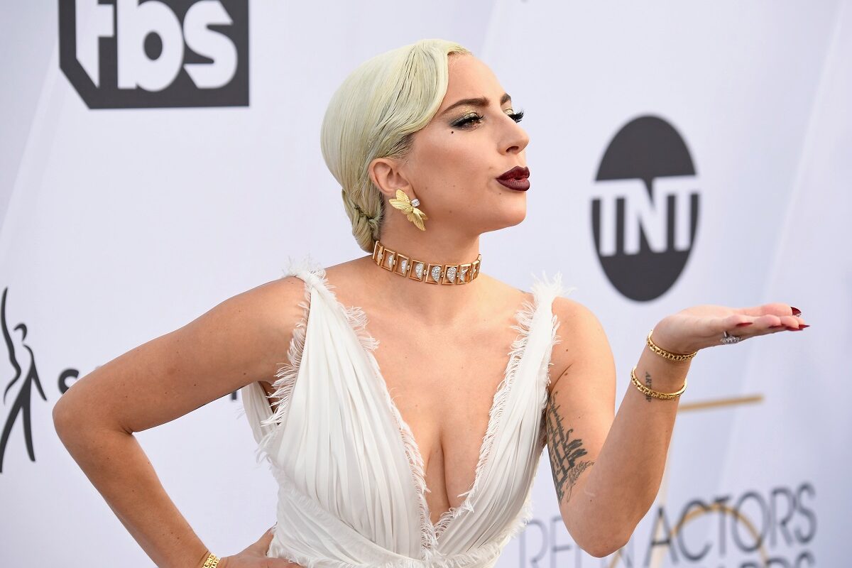 Lady Gaga la Screen Actors Guild Awards, ediția 25, 2019. Pe covorul roșu, într-o rochie albă, cu decolteu, bijuterii de aur și ruj roșu închis, fundal alb