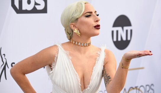 Lady Gaga la Screen Actors Guild Awards, ediția 25, 2019. Pe covorul roșu, într-o rochie albă, cu decolteu, bijuterii de aur și ruj roșu închis, fundal alb