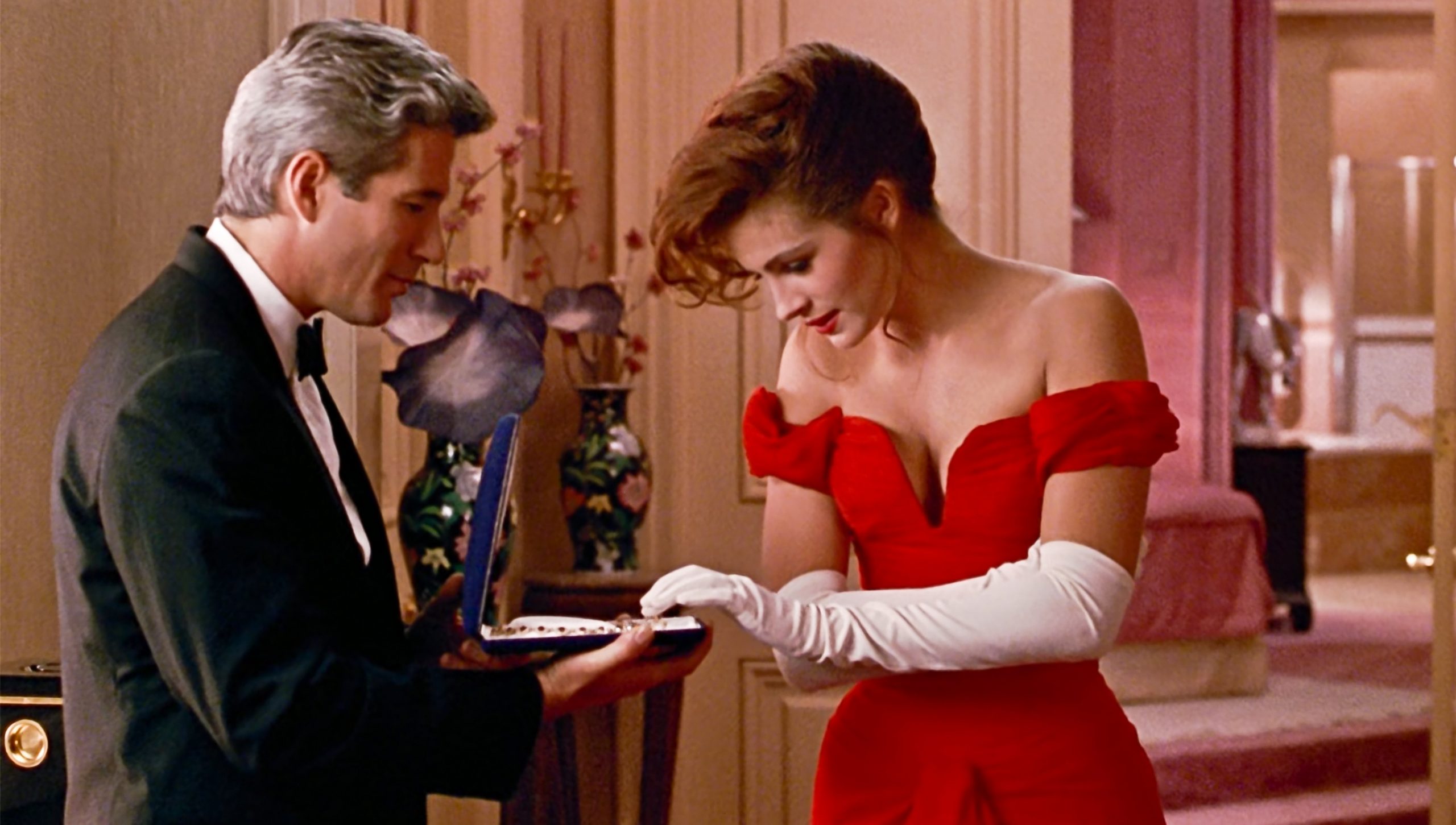 Julia Roberts și Richard Gere în filmul Pretty Woman, din 1990. Scena în care el îi oferă colierul. Gere e imbrăcat în costum negru, Julia poartă rochia roșie decoltată și mănuși albe, în camera de hotel