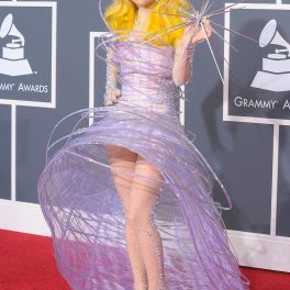 Lady Gaga pe covorul roșu la Premiile Grammy, cea de-a 52-a ediție. A purtat o rochie mov, cu cercuri mari, părul foarte blond și pantofi înalți care sclipesc. Fundal roșu și gri