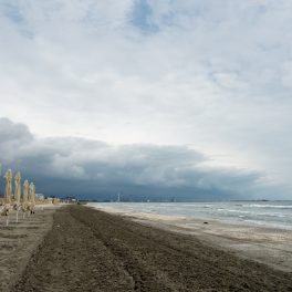Un peisaj superb cu plaja din Năvodari pe care se află umbreluțe, în timp ce plaja este lovită de valurile mării, iar cerul este înnorat