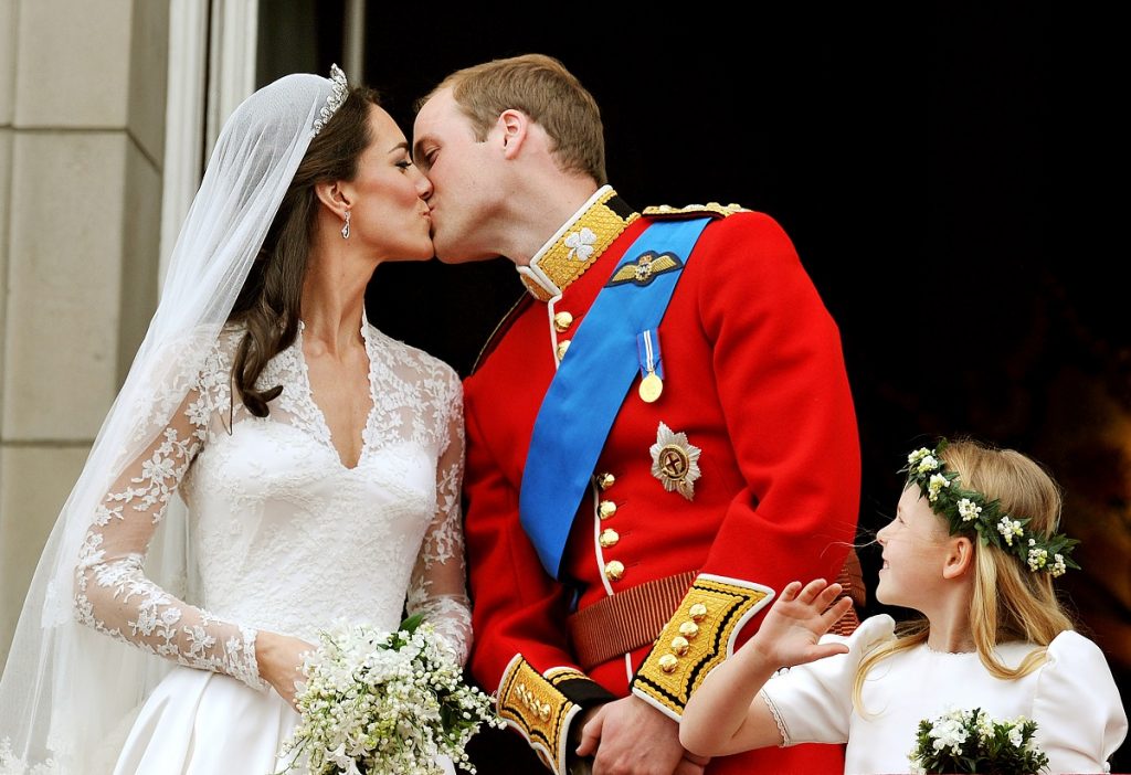 Kate Middleton și prințul William în ziua nunții din anul 2011, sărutul pe balcon după ceremonei. Ea în rochie de mireasă, el în uniforma roșie.