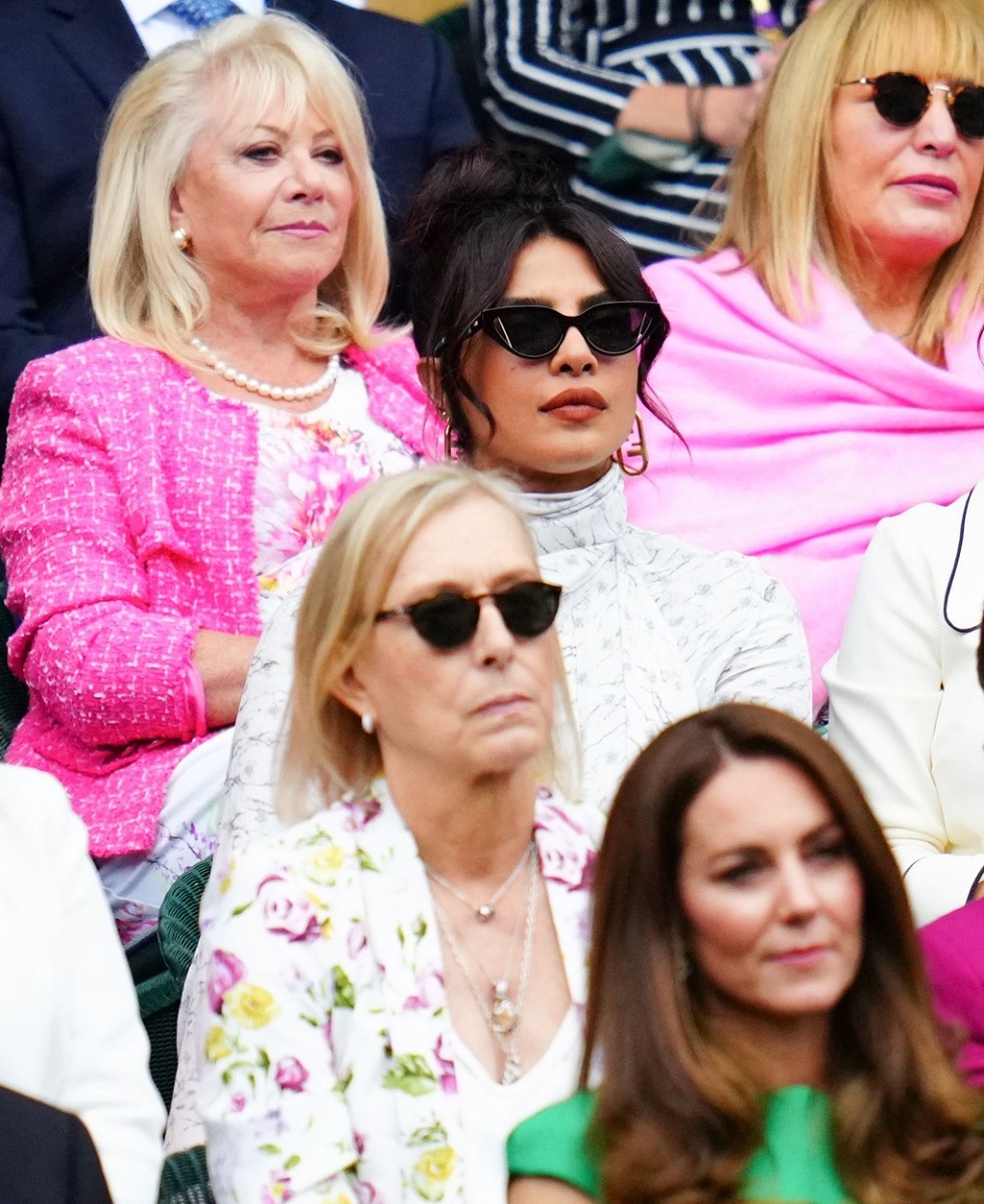 Priyanka Chopra în tribunele Turneului de la Wimbledon în iulie 2021, imbracata în rochie albă, ochelari de soare, părul prins sus. În spatele ei, două femei îmbrăcate în roz