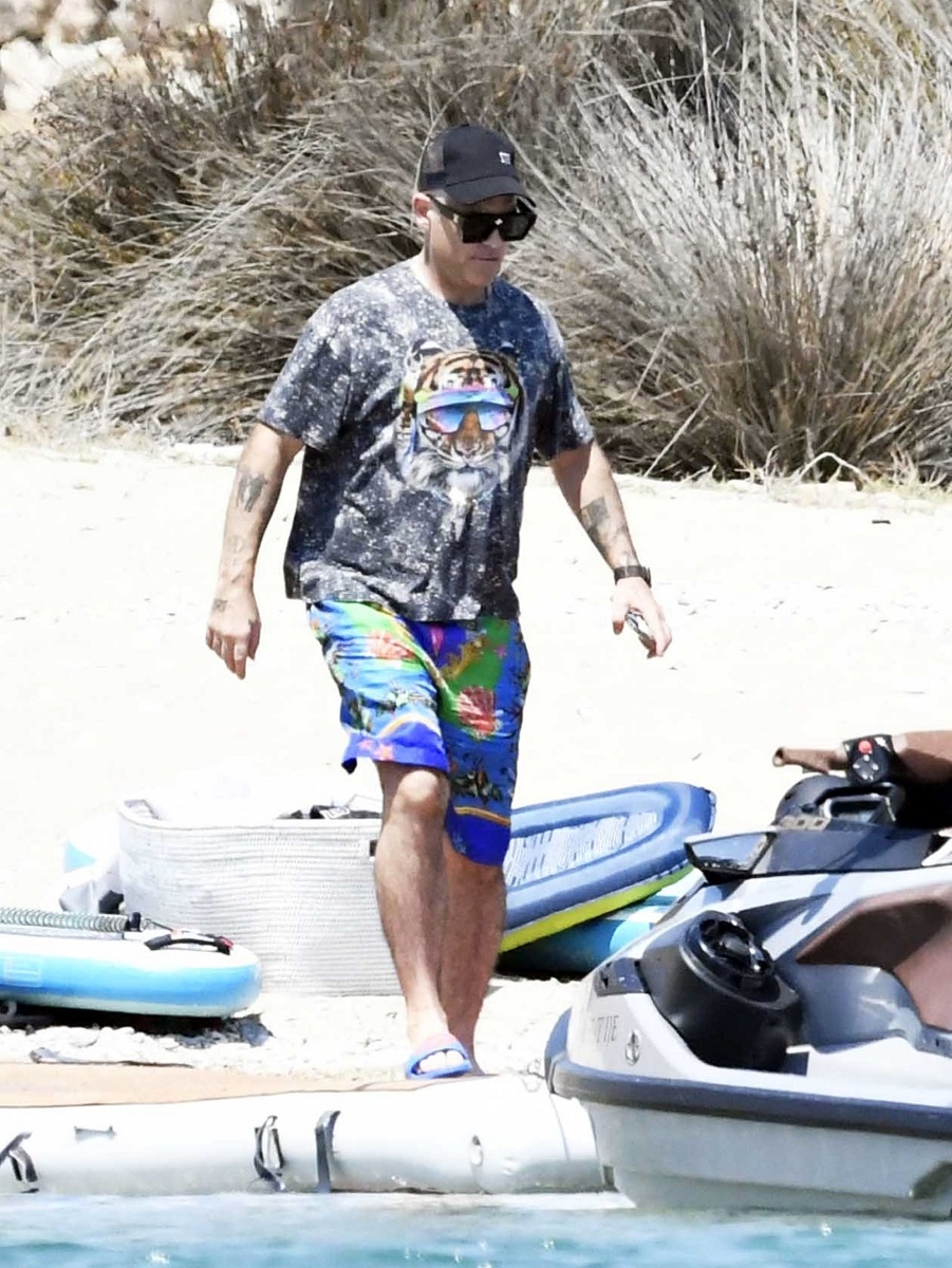 Robbie Williams a fost surprins pe plaja din Grecia, îmbrăcat în pantaloni scurți de culoare albastră, tricou gri și șapcă neagră. Fundal cu plajă