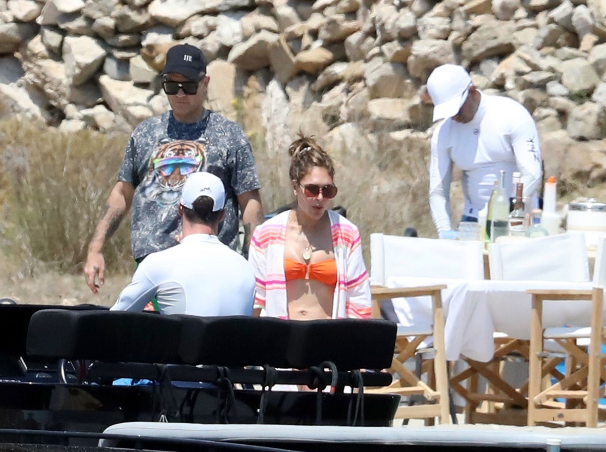 Robbie Williams și Ayda Field pe plajă în grecia, se întorc peo barcă neagră. Doi angajați îmbrăcați în alb, plaja în spate, Robbie Williams îmbrăcat în tricou gri, Ayda îmbrăcată în costum de baie portocaliu