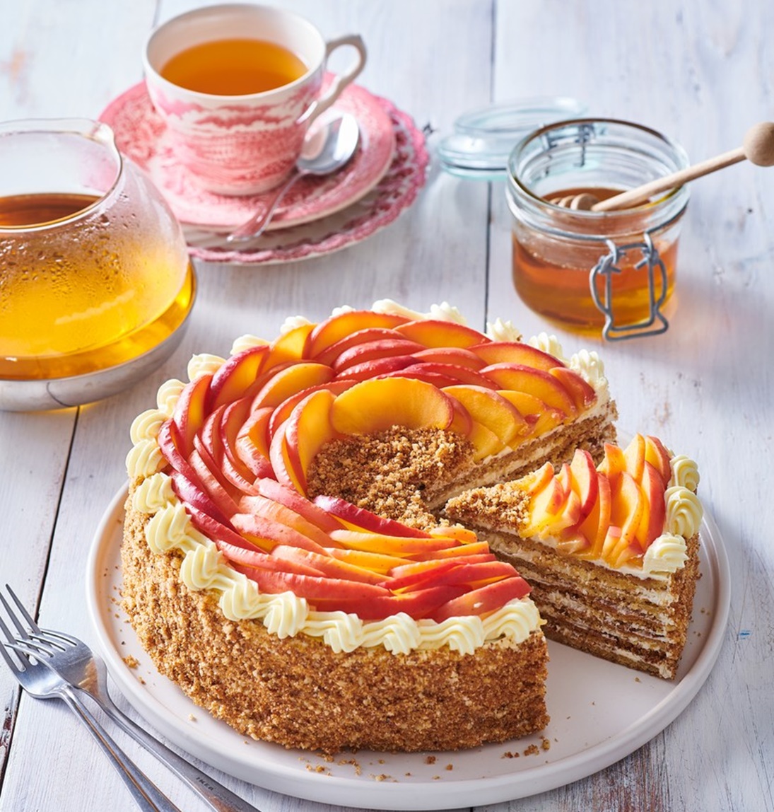 Tort Medovik secționat pe un platou alb, alături de cești cu ceai și un borcan cu miere