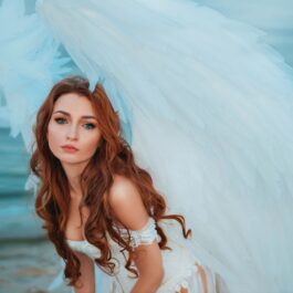 O femeie frumoasă cu părul lung și roșcat care poartă o rochie albă și are înspate aripi de înger în timp ce stă în genunchi pe o plajă reprezentând una dintre cele mai sincere zodii