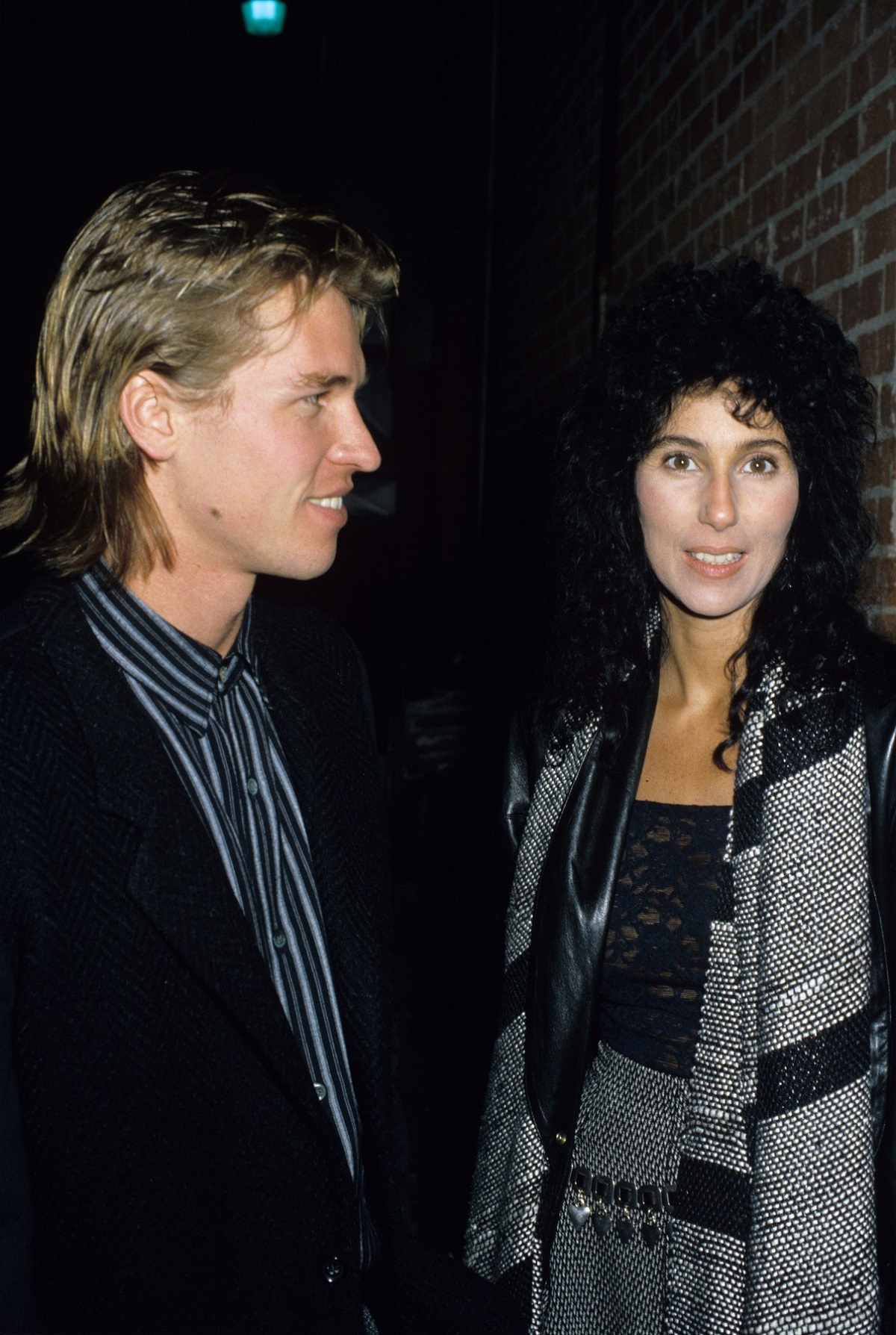 Cher care poartă o bluză neagră din dantelă și o fustă gri, alături de iubitul său Val Kilmer în anul 1984