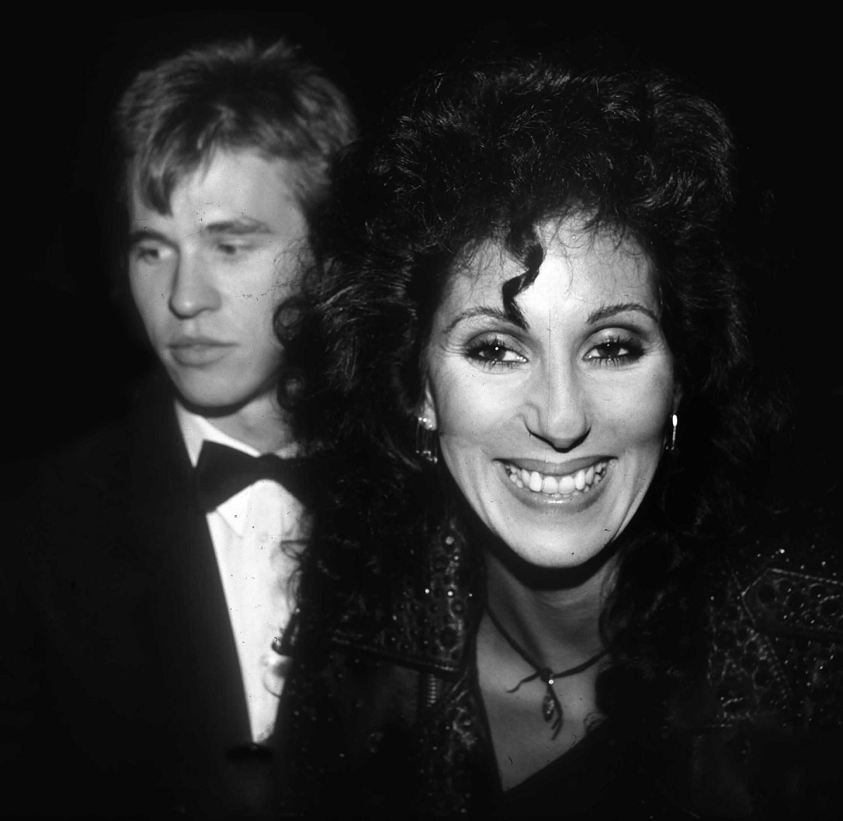 Cântăreața Cher zâmbind, alături de fostul său iubit, Val Kilmer, care poartă un costum cu papion în anul 1987