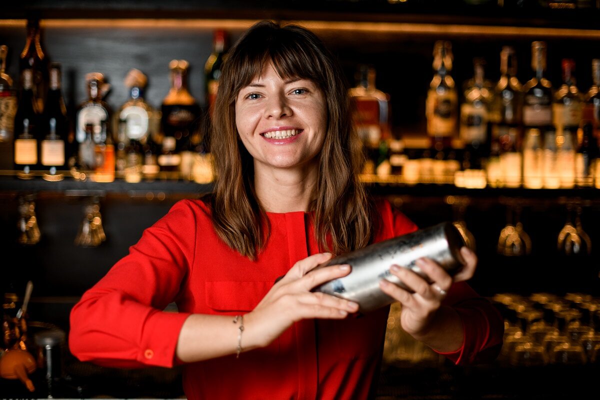 O femeie frumoasă care poartă o bluză roșie și prepară un cocktail cosmopolitan și zâmbește în timp ce se află în spatele unui bar