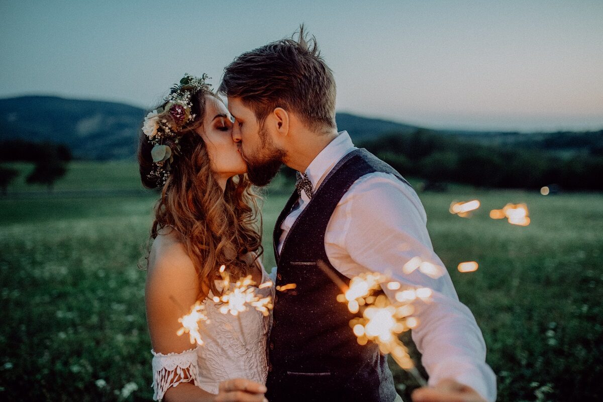 Un cuplu de îndrăgostiți în timp ce se sărută și țin în mână artificii pe un câmp liber
