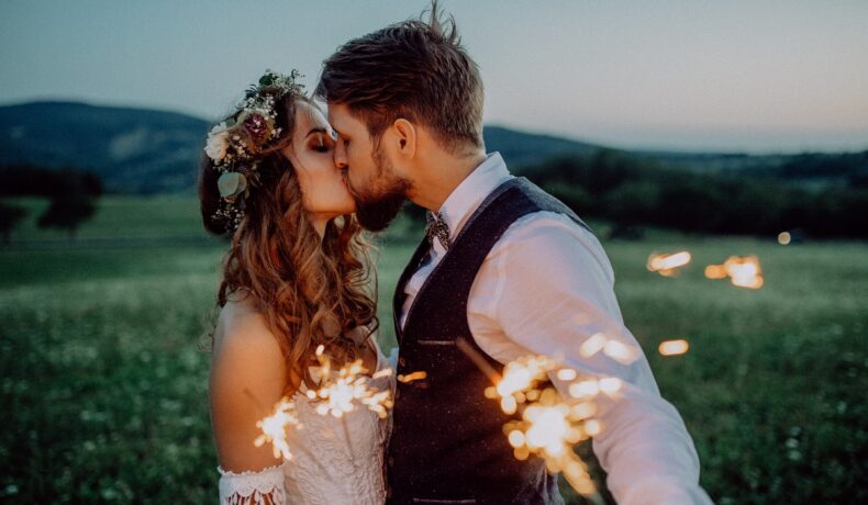 Un cuplu de îndrăgostiți în timp ce se sărută și țin în mână artificii pe un câmp liber
