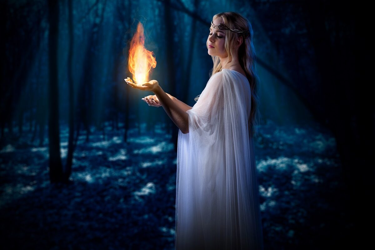 O femeie frumoasă într-o rochie albă care stă într-o pădure întunecată în tim ce ține în mână o flacără