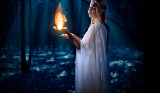 O femeie frumoasă într-o rochie albă care stă într-o pădure întunecată în tim ce ține în mână o flacără