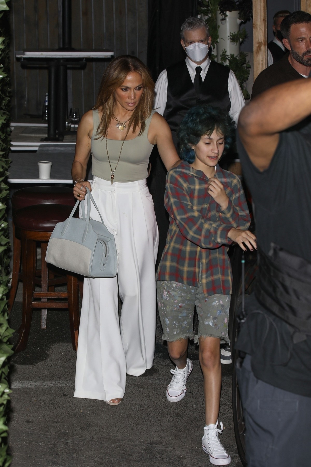 Jennifer Lopez într-o pereche de pantaloni albi și un top crem în timp ce ține în mână o poșetă și o conduce pe fiica sa Emme la mașina familiei în Los Angeles