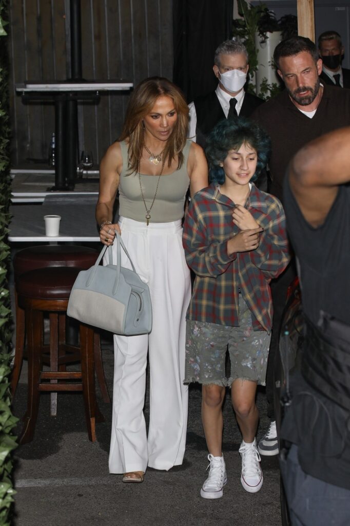 Jennifer Lopez purtând un top gri în timp ce poartă o pereche de pantaloni albi și ține în mână o poșetă și o însoțește pe fiica sa Emme la mașină, unde se află și Ben Affleck