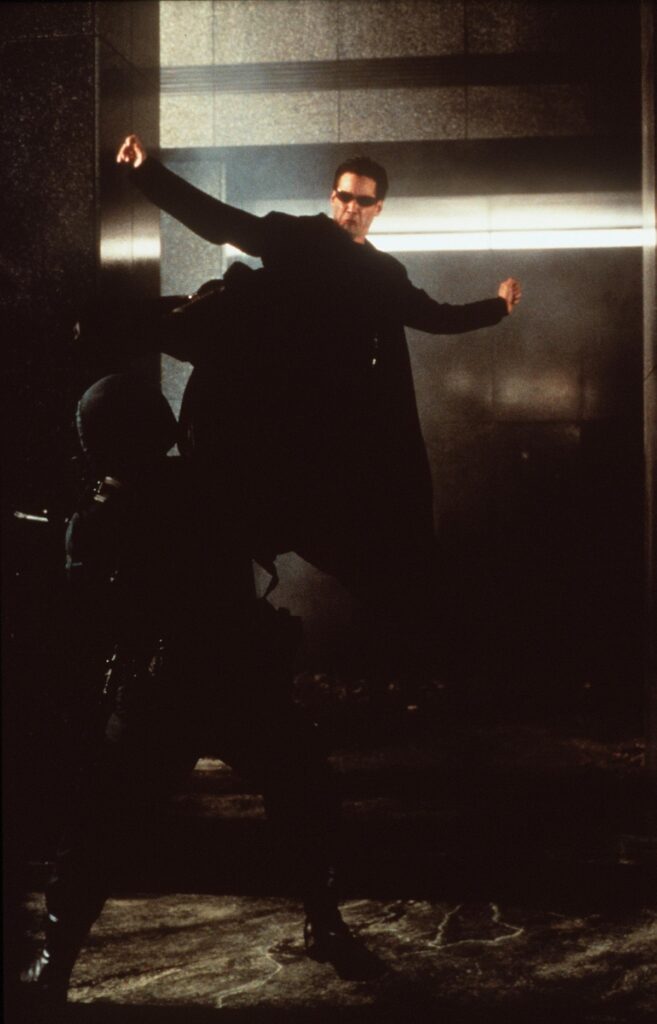 Actorul Keanu Reeves într-un costum negru în timp ce sare în aer și lovește cu piciorul un bărbat într-o scenă din filmul Matrix