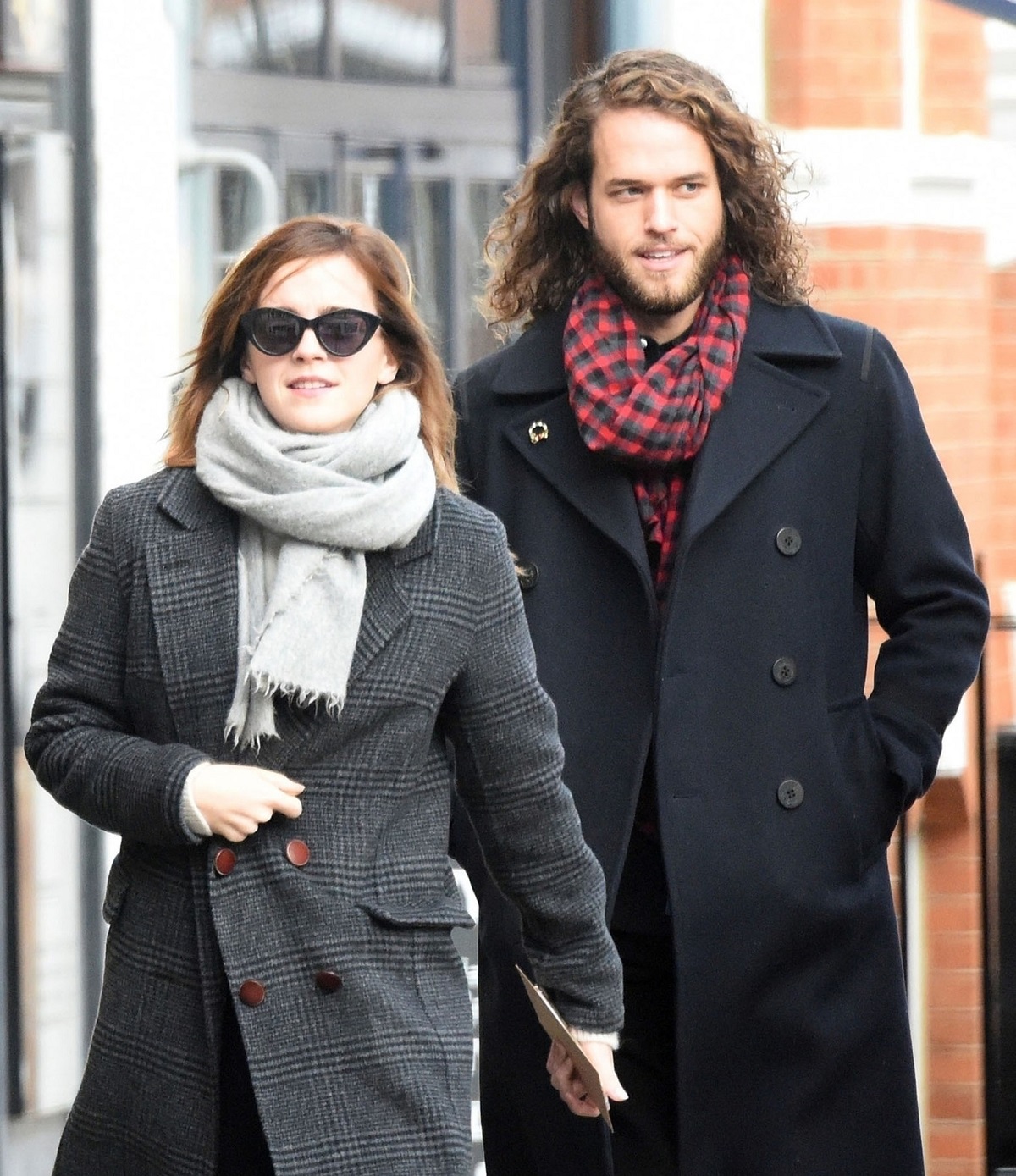 Emma Watson purtând un palton gri, un fular și o pereche de ochelari de soare în timp ce merge pe stradă alături de iubitul său, Leo Robinton care poartă un palton negru
